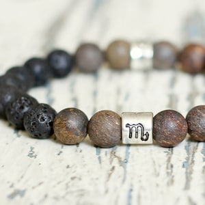 Op de afbeelding: Een kralenarmband met zwarte lavasteenkralen, bruine stenen kralen en een zilverkleurige spacer kraal met een Schorpioen symbool. De armband is geregen op een elastisch koord.