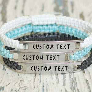 Könnte beinhalten: Drei übereinanderliegende Armbänder mit verstellbaren Metalletiketten, die mit individuellem Text personalisiert werden können. Die Armbänder sind aus weißem, schwarzem und hellblauem Band gefertigt.