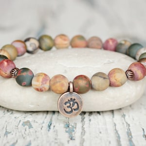 May include: A multi-colored stone bracelet with a copper Om charm. The bracelet is made of round, flat stones in shades of brown, red, green, and yellow. The Om charm is a round disc with the Sanskrit symbol for Om.