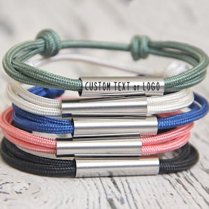 Puede incluir: Una pila de cinco pulseras de cuerda de colores vivos con barras de metal plateado que se pueden personalizar con texto o un logotipo.