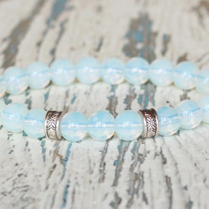 Op de afbeelding: Een armband met opalescente, ronde kralen. De kralen zijn zacht wit met een subtiele iriserende glans. Zilverkleurige, patroon spacers zijn tussen sommige kralen geplaatst. De armband wordt getoond op een verweerd, lichtgeleurd houten oppervlak.