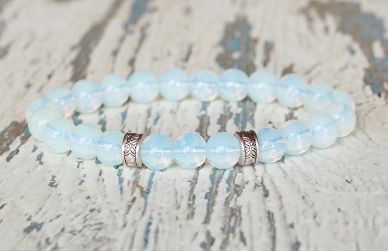 Op de afbeelding: Een armband met opalescente, ronde kralen. De kralen zijn zacht wit met een subtiele iriserende glans. Zilverkleurige, patroon spacers zijn tussen sommige kralen geplaatst. De armband wordt getoond op een verweerd, lichtgeleurd houten oppervlak.