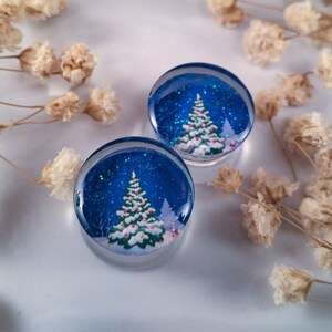 May include: Two round, clear glass ornaments with a blue background and a white, snow-covered Christmas tree with red ornaments. The ornaments are filled with glitter.