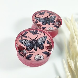 Op de afbeelding: Een paar roze acryl oorpluggen met een bloemen- en vlinderontwerp. De pluggen hebben een zwart-witte vlinder met een roze, witte en zwarte bloemenprint.