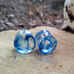 May include: A pair of clear glass ear gauges with blue swirls inside. The gauges are on a light brown surface.