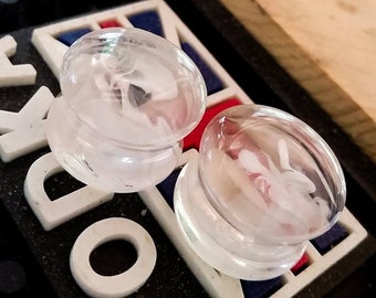 May include: Two clear glass ear gauges with a white swirl design. The gauges are on a black surface with white lettering.