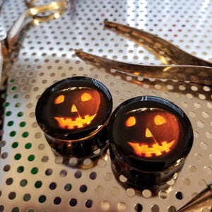 Puede incluir: Dos tapones para orejas de Halloween de color negro y naranja con un diseño de cara de calabaza.