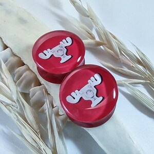 Puede incluir: Dos expansores de oreja circulares rojos con un diseño de alce blanco. Los expansores están hechos de un material brillante y se muestran sobre una superficie blanca con plantas secas. Un accesorio único.