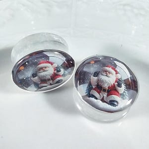 May include: Pair of clear glass Christmas themed ear gauges with a Santa Claus image inside. The Santa Claus is wearing a red suit with white trim and a white beard. The background is a snowy scene.