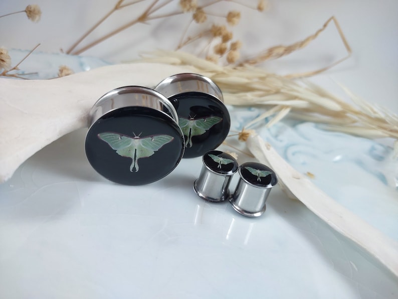 May include: A set of three silver ear gauges with a black background and a green luna moth design.