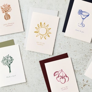 May include: Set of ten greeting cards with various illustrations and text. The cards feature a bouquet of flowers, a sun, a cocktail, a bunch of cherries, and a bouquet of flowers. The text on the cards includes "You're blooming wonderful", "You are my sunshine", "Cheers to you", "You're the sweetest", and "Thanks a bunch".