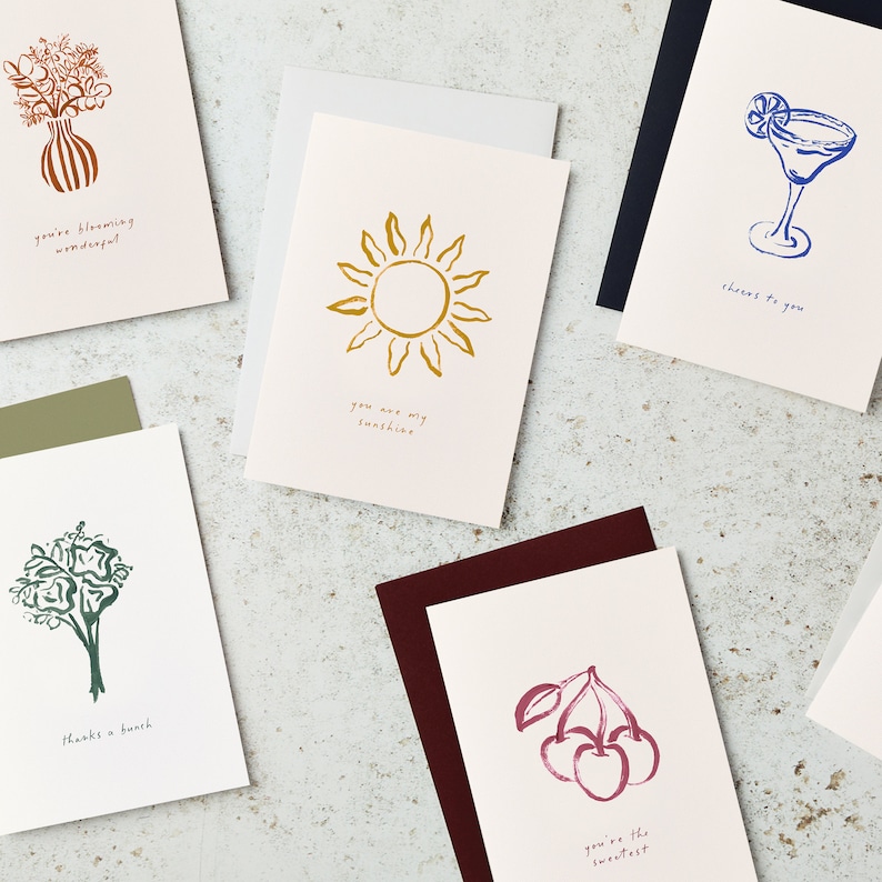 May include: Set of ten greeting cards with various illustrations and text. The cards feature a bouquet of flowers, a sun, a cocktail, a bunch of cherries, and a bouquet of flowers. The text on the cards includes "You're blooming wonderful", "You are my sunshine", "Cheers to you", "You're the sweetest", and "Thanks a bunch".
