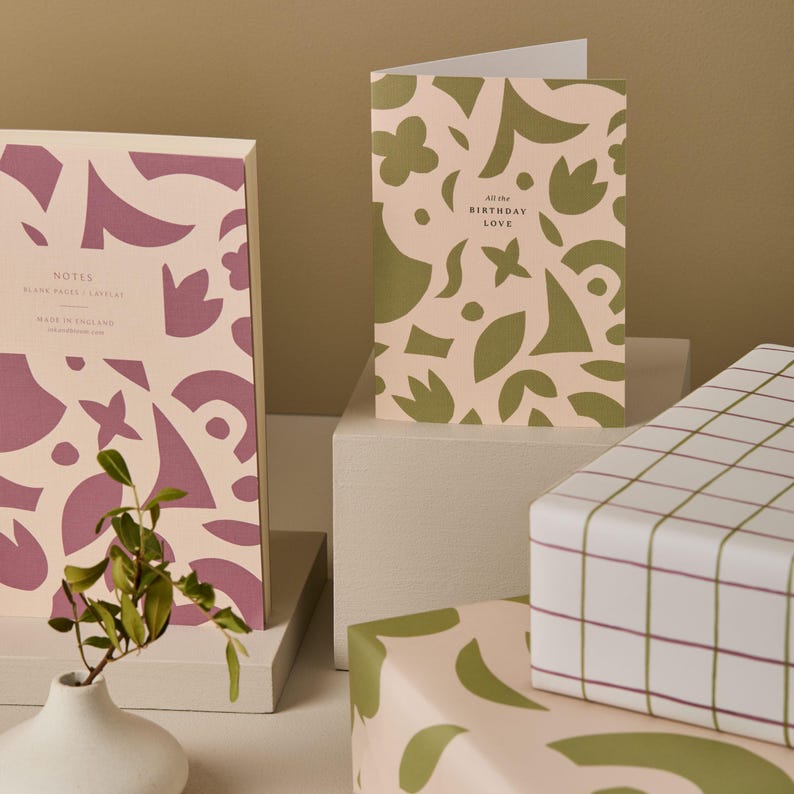 May include: A stationery set featuring a notebook, greeting card, and wrapped gift boxes. The notebook and card have a pink background with purple and green abstract shapes. The card reads "All the Birthday Love". The gift boxes are wrapped in patterned paper.