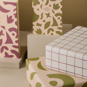 May include: Gift boxes and greeting cards with abstract designs. The boxes are wrapped in patterned paper with green and burgundy accents. One card reads "All the Birthday Love". The overall color scheme is soft pink and white.