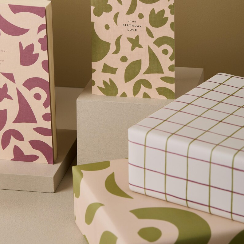 May include: Gift boxes and greeting cards with abstract designs. The boxes are wrapped in patterned paper with green and burgundy accents. One card reads "All the Birthday Love". The overall color scheme is soft pink and white.