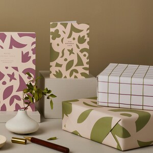 May include: A still life arrangement featuring stationery and gift wrap. A notebook and greeting card display a floral pattern in mauve and olive green. Gift wrap with a grid and floral design is also present.