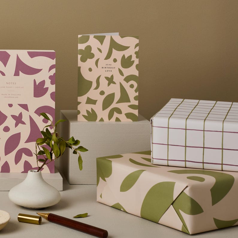 May include: A still life arrangement featuring stationery and gift wrap. A notebook and greeting card display a floral pattern in mauve and olive green. Gift wrap with a grid and floral design is also present.