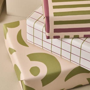 May include: Three stacked gift boxes wrapped in patterned paper. The top box has a striped design in olive green and burgundy on a cream background. The middle box has a grid pattern in burgundy and olive green on a white background. The bottom box has an abstract olive green design on a cream background.