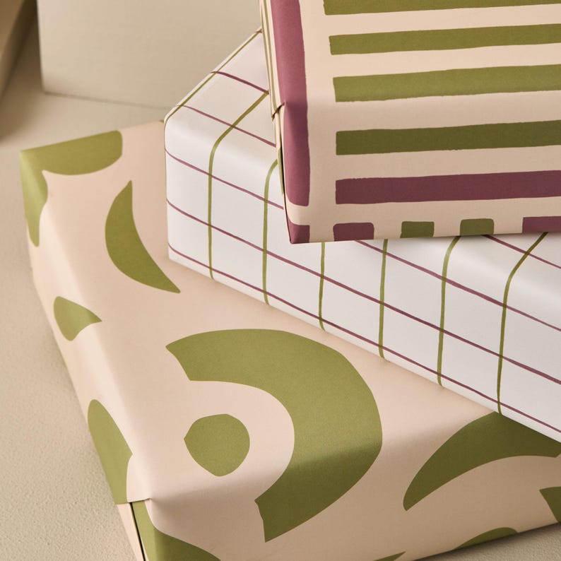 May include: Three stacked gift boxes wrapped in patterned paper. The top box has a striped design in olive green and burgundy on a cream background. The middle box has a grid pattern in burgundy and olive green on a white background. The bottom box has an abstract olive green design on a cream background.