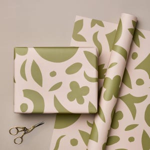 May include: A wrapped gift box and rolls of wrapping paper with a repeating pattern of olive green abstract shapes on a pale pink background. A pair of gold scissors is next to the gift box.