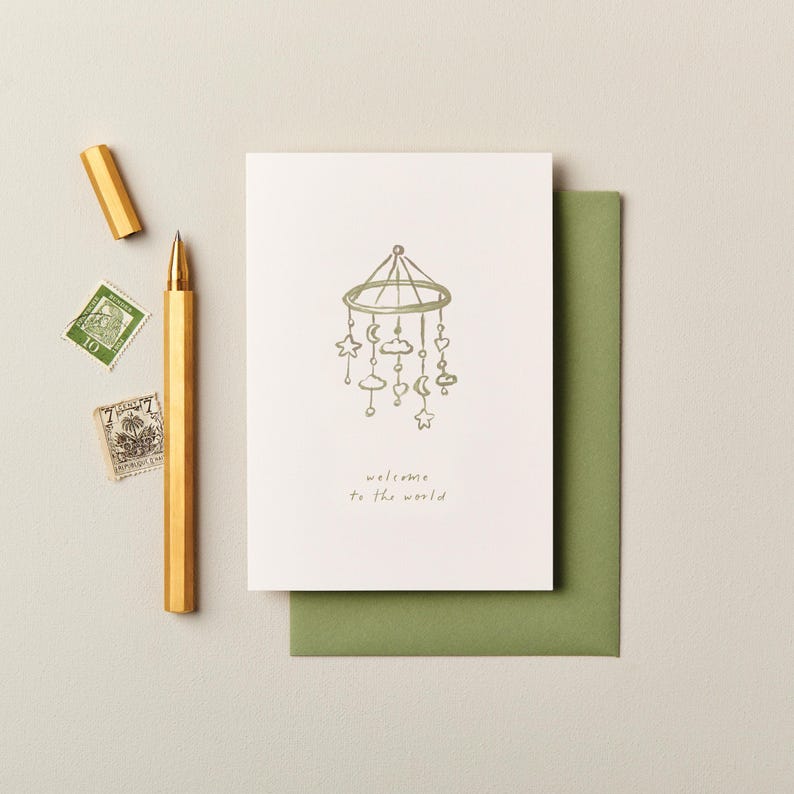 K&ouml;nnte beinhalten: Eine Gru&szlig;karte mit einem Baby-Mobile-Design in olivgr&uuml;ner Tinte und dem Schriftzug "welcome to the world". Die Karte liegt auf einem gr&uuml;nen Umschlag. Ein goldener Stift und zwei Vintage-Briefmarken sind ebenfalls im Bild.