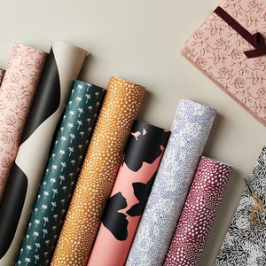 May include: An assortment of patterned wrapping paper rolls in various designs, including floral, palm tree, and abstract patterns. A wrapped gift box with a burgundy ribbon and a sheet of wrapping paper with scissors are also visible.