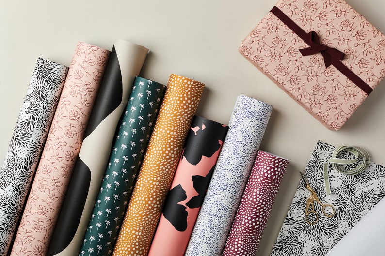 May include: An assortment of patterned wrapping paper rolls in various designs, including floral, palm tree, and abstract patterns. A wrapped gift box with a burgundy ribbon and a sheet of wrapping paper with scissors are also visible.