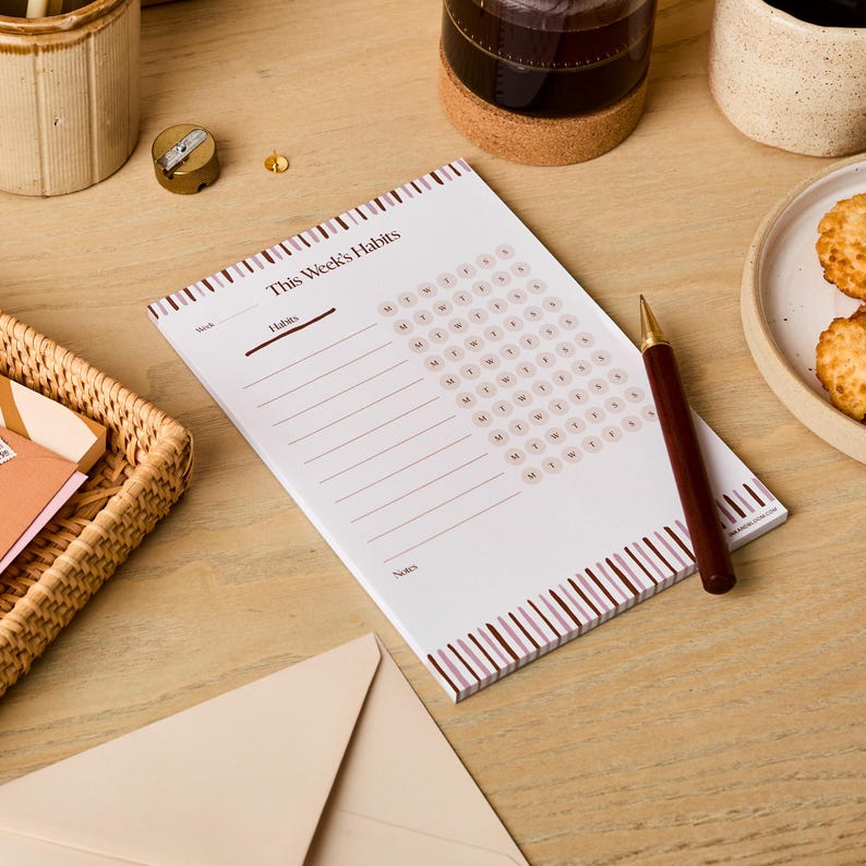May include: A white notepad with brown stripes on the edges. The notepad has a title that says "This Week's Habits". There is a section for listing habits and a grid for tracking habits throughout the week. The notepad is on a wooden surface with a pen and other items.