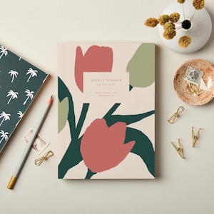 May include: A weekly planner with a floral design in red, green, and beige. The planner has the text "WEEKLY PLANNER ANY DATE DIARY MADE IN ENGLAND". A pencil, stamps, and paper clips are also visible.