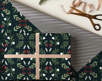 May include: A wrapped gift with a dark green floral pattern, tied with a tan ribbon. The wrapping paper features white flowers, red berries, and green leaves. A striped paper and scissors with a ribbon are in the background.