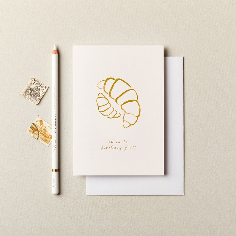 May include: A white card with a gold outline of a croissant and the text "oh la la birthday girl!"