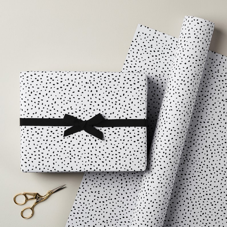 May include: A white gift wrap with a black polka dot pattern. A black ribbon is tied around a wrapped gift box. A pair of gold scissors is on the table.