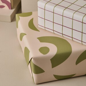 May include: Two wrapped gift boxes. One is covered in light pink paper with large olive green abstract shapes. The other box is wrapped in white paper with a grid pattern of olive green and burgundy lines.