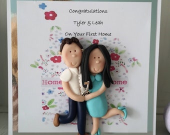 May include: A handmade greeting card with the text "Congratulations Tyler & Leah On Your First Home." The card features a clay sculpture of a couple holding a key, with a floral house design in the background.