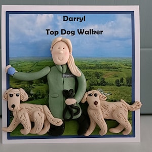 Könnte beinhalten: Eine Grußkarte mit einer 3D-Tonfigur einer Person in einem grünen Mantel, die eine schwarze Leine und einen blauen Ball hält, flankiert von zwei hellen Hunden. Der Hintergrund zeigt eine grüne Landschaft unter blauem Himmel mit dem Text Darryl Top Dog Walker.
