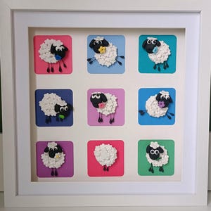 Puede incluir: Una obra de arte enmarcada con nueve paneles cuadrados, cada uno mostrando un diseño de oveja. Las ovejas tienen lana blanca, caras negras y están sobre fondos coloridos de rosa, azul, morado y verde. La obra está presentada en un marco blanco.