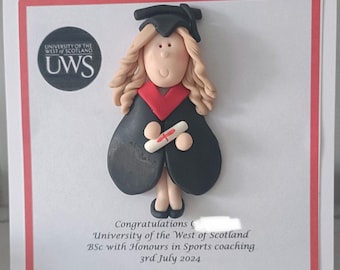 May include: A graduation card featuring a handmade figurine of a graduate in a black cap and gown with a red collar, holding a diploma. The card includes the University of the West of Scotland logo and congratulatory text.