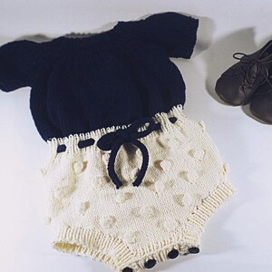Peut inclure: Un barboteuse pour bébé tricotée bleu marine et crème avec un cordon de serrage et des détails texturés. La barboteuse est associée à des chaussures de bébé en cuir marron. La tenue est conçue pour les nourrissons.