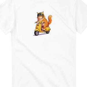 May include: White t-shirt featuring a cartoon illustration of an orange cat riding a yellow scooter. The cat wears goggles and a checkered helmet. The scooter has black tires and a yellow seat.