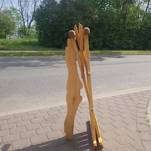 May include: A wooden easel with a natural finish and rounded wooden knobs at the top. The easel is folded and standing on a brick walkway.
