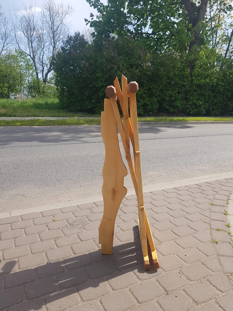 May include: A wooden easel with a natural finish and rounded wooden knobs at the top. The easel is folded and standing on a brick walkway.