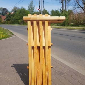 May include: A wooden folding chair with a natural finish. The chair is folded up and standing on a sidewalk.
