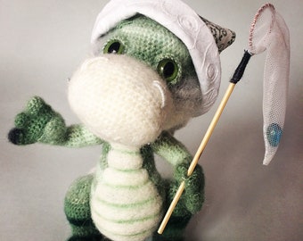 Peut inclure: Une figurine de dragon crochetée verte avec un ventre et un museau blancs, portant un chapeau blanc à motif subtil. Le dragon a de grands yeux verts et tient un petit bâton en bois avec un filet en maille blanche contenant une bille bleue.