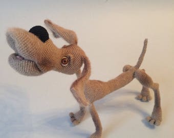 Puede incluir: Una figura de perro tejida a ganchillo en color beige con un cuerpo largo y esbelto, patas delgadas y orejas caídas. El perro presenta una nariz negra prominente, una boca muy abierta y un ojo marrón visible. La cola apunta hacia arriba.