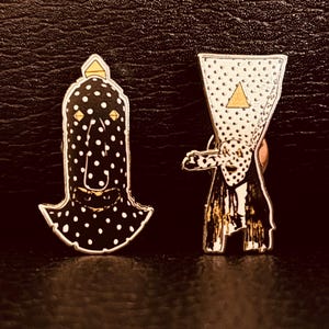 May include: Two enamel pins with unique designs. One pin is a black figure with white polka dots and a gold triangle on top. The other pin is a white figure with black polka dots, a gold triangle, and a gold-colored base.