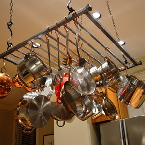 May include: A metal pot rack with multiple hooks hanging from the ceiling. The rack is filled with various sizes of stainless steel pots and pans, including a copper pot.