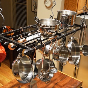 May include: A black metal pot rack with multiple hooks hanging from the ceiling. The rack is filled with various sizes of pots, pans, and kitchen utensils. The rack is hanging over a wooden countertop.