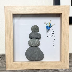May include: A framed artwork featuring a stack of grey stones with a blue and white origami crane flying above them. The crane is flying in a dotted line path.