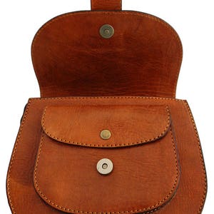 May include: A brown leather saddlebag with a flap closure and a small front pocket. The bag has a rounded shape and a handle on top. The bag is made of a smooth, textured leather and has a magnetic snap closure.