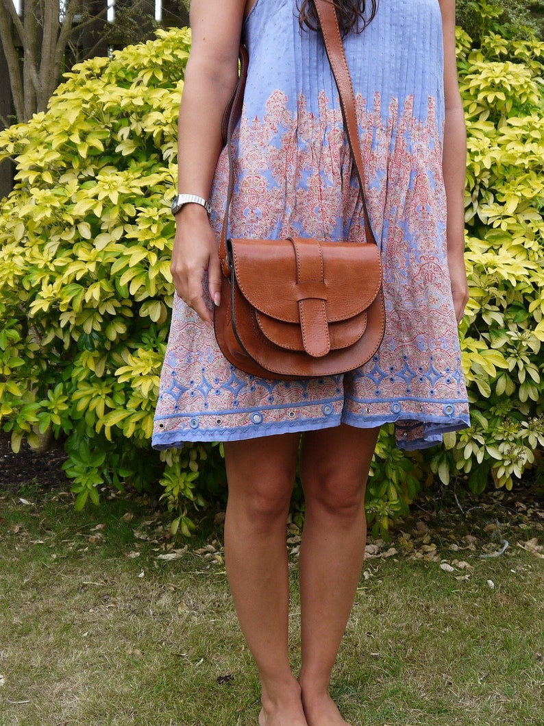 May include: A brown leather saddlebag with a long strap is worn over the shoulder. The bag has a curved shape with a flap closure. The person is wearing a blue and red patterned dress. The background features green foliage and grass.